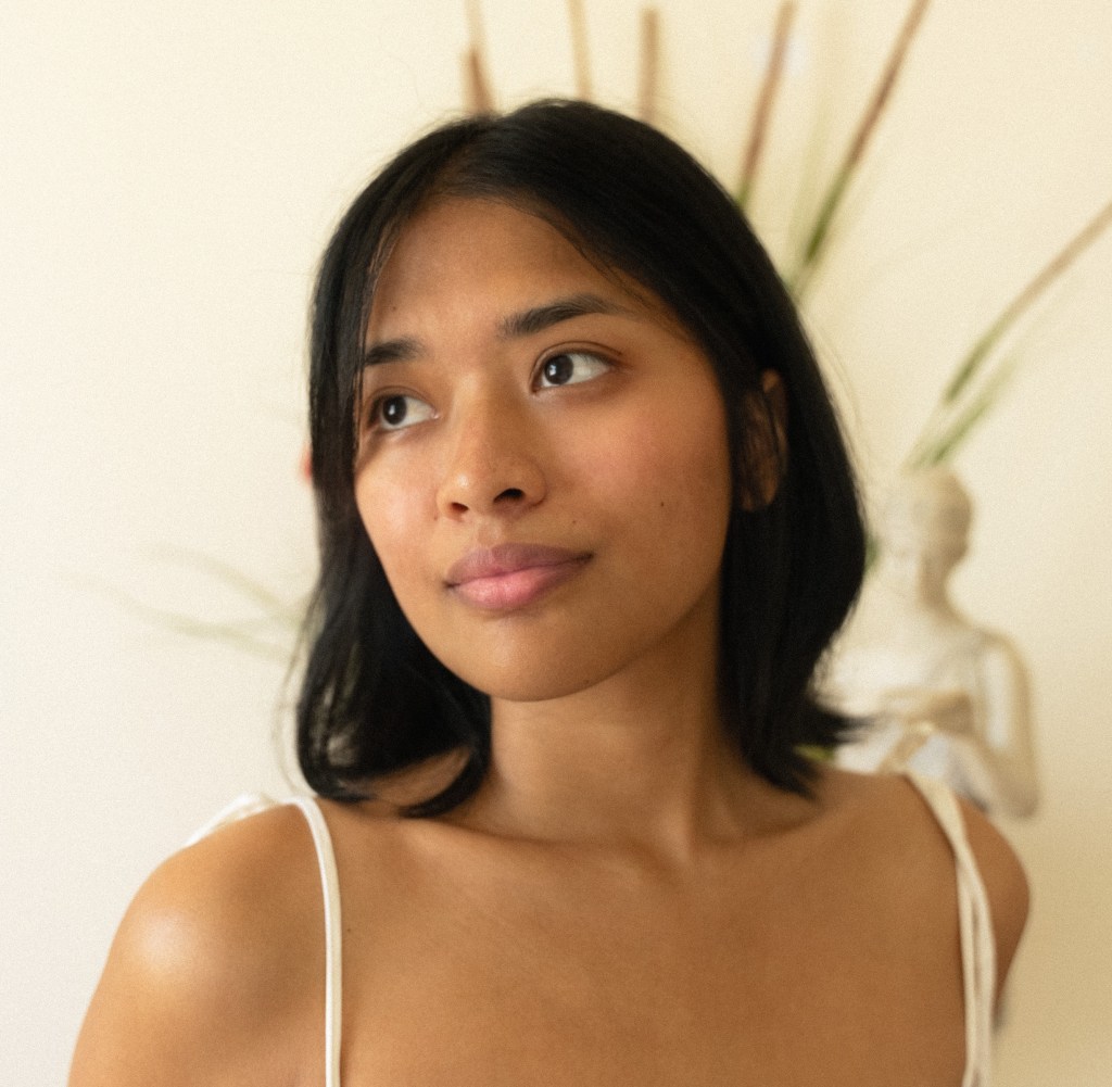 A warm-toned portrait of Kianna Reyes looking away from the camera to her right with a subtle smile. She has straight, dark shoulder-length hair with middle-parted bangs and is wearing a white top with thin shoulder straps. In the softly blurred background, there is a neutral off-white wall with dried tall grass stalks and a small decorative figurine.