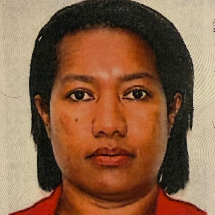 Passport-style photo of Kila Wani looking directly at the camera with a neutral expression. She has short, shoulder-length dark hair pushed back behind her ears and is wearing a red blazer or jacket over a dark top against a light gray or off-white background.