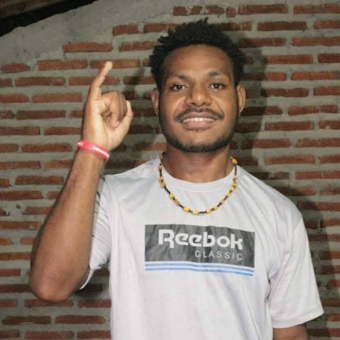 Medium portrait of Oskar Ugipa standing indoors in front of an unpainted red brick wall. He is smiling at the camera while raising his right index finger upward. He has short curly hair, a light beard and mustache, and is wearing a white t-shirt with the logo "Reebok Classic," a black and yellow beaded necklace, and a red band on his wrist.